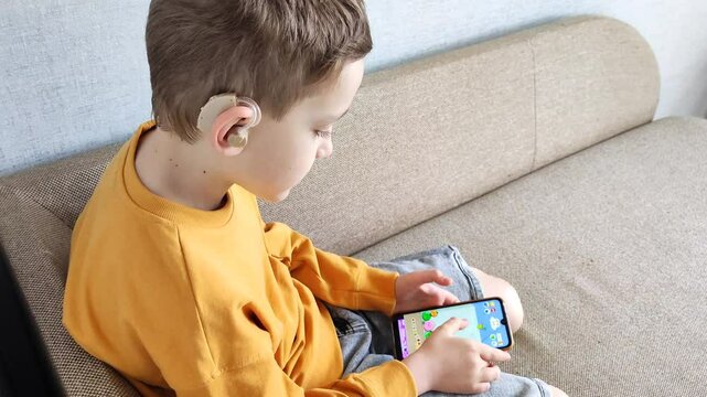 Boy with hearing aid in ear holds phone in hands, listens to audiobook while sitting in his room