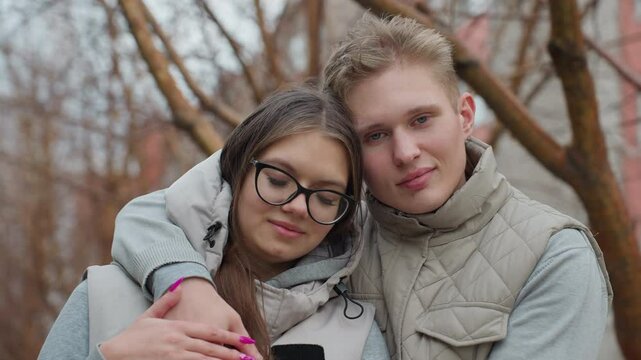 Beautiful moment of lovely couple holding each other warmly in cozy outdoor setting, standing close together with gentle expressions as trees without leaves sway behind them