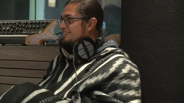 Medium shot of a young man with glasses and a beard, sitting casually in a professional recording studio environment. He wears headphones around his neck and a distinctive patterned poncho or sweater.