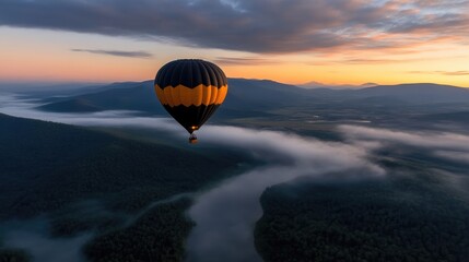 Obraz premium Hot air balloon above misty mountains at sunrise. A breathtaking aerial view of a black and yellow hot air balloon soaring through a valley shrouded in morning mist.