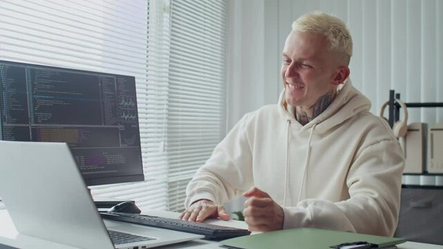 Medium shot of tattooed male specialist feeling happy while working on coding scripts at desk in office