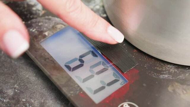Close up of woman hand with manicured nails pressing tare button on digital kitchen scale while metal container rests on surface, resetting measurement to zero for accurate weighing
