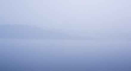 Fototapeta premium Serene fog-covered lake at dawn with distant mountains and soft reflections