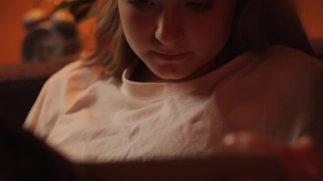 Relaxed girl read digital book on tablet, sitting comfortably in soft evening illumination, wearing white t-shirt