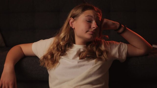 Sleepy blonde female lounging on dark couch, stretching lazily and yawning, embodying morning drowsiness in comfortable home setting