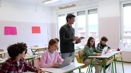 Male teacher holding tablet and walking around classroom while students are using laptops and taking notes