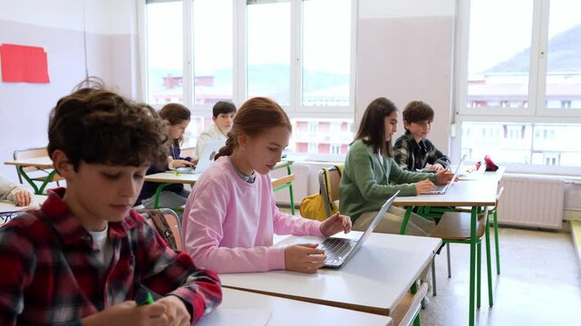 Middle school students learning and working on laptops and notebooks in a classroom, supervised by their teacher