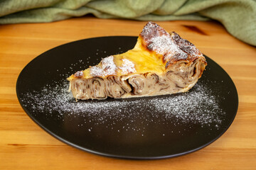 Oven baked rolled pancakes pie slice sprinkle with powder sugar, on a black plate on wooden table with   green curtains
