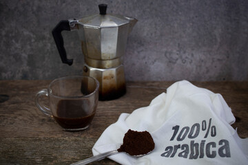 100% arabica written with stamp letters on a white fabric sack next to a mocha cup and a spoon full of ground coffee, soft focus close up