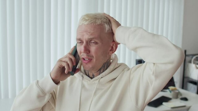 Medium close up of anxious male tattooed Caucasian developer calling on smartphone at office