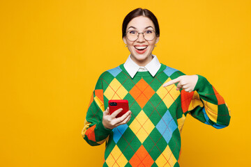 Young amazed woman she wear glasses green sweater casual clothes hold in hand use point index finger on mobile cell phone isolated on plain orange yellow background studio portrait. Lifestyle concept.