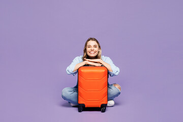 Traveler smiling woman wear blue clothes hold suitcase bag sit look camera isolated on plain purple...