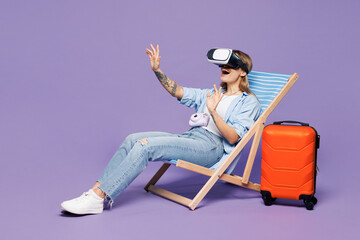Traveler woman wears blue clothes glasses sit in deck chair watch in vr headset pc gadget isolated on plain purple background. Tourist travel abroad in free time rest getaway. Air flight trip concept.