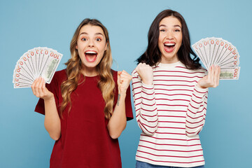 Young winner shocked couple two friends women wearing red casual clothes together hold in hand fan of cash money in dollar banknotes isolated on pastel plain light blue background. Lifestyle concept.