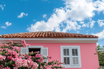 Charming pink house with floral walls, red-tiled roof, and white shutters under a bright blue sky with fluffy clouds 