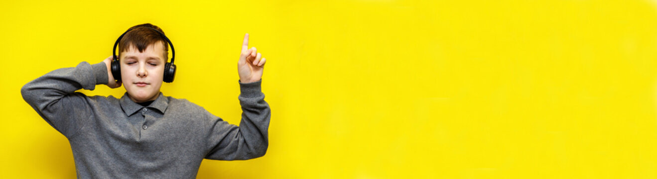 ten year old boy in grey knitted sweater enthusiastically listening to music in black headphones on yellow background. closed eyes and raised index finger up
