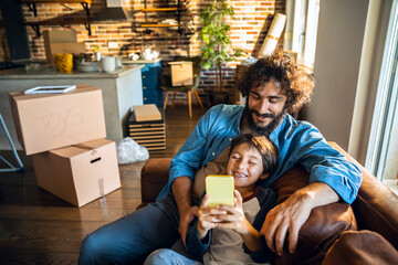 Father and son relaxing on couch and watching videos on smartphone after moving into new home