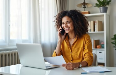 Obraz premium Happy woman talks on cellphone, smiles, holds paper documents at desk. Uses laptop computer for online work. Freelance job, remote work, business career, entrepreneurship, online communication.