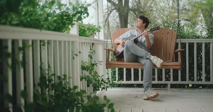 Young male musician sitting on a porch, playing guitar and writing a song