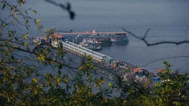 A view from the top of a mountain offers a perspective of Elephanta Island and its boat jetty. Several tourist catamaran boats are docked nearby.
