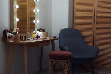 Makeup room. Mirror, wooden dressing table, different beauty products, stool and armchair indoors