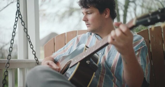 Young male musician sitting on a porch, playing guitar and writing a song