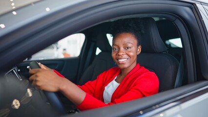 Adorable African American woman sitting in luxury car. Satisfied girl holding hands on steering wheel while imagining driving. Positively looking at camera and cheerfully smiling after test drive.