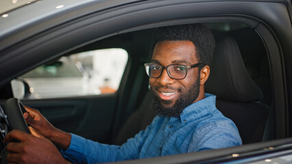 Side view of African American man sitting in car. Happy male holding hands on steering wheel and positively looking directly at camera. Getting ready for driving new car or starting test drive.