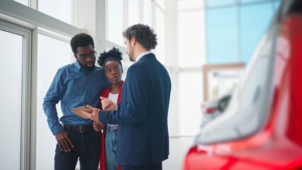 Attractive manager offering to get acquainted with new cars on tablet device. Couple listening with...