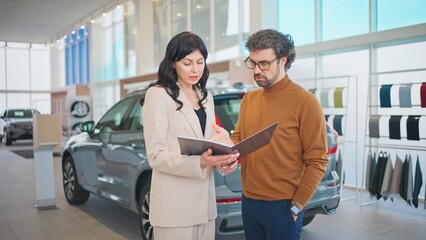 Obraz premium Camera view of Caucasian beautiful woman in business suit showing variants or car models in catalogue to her potential buyer. People talking together while standing in middle of auto salon.