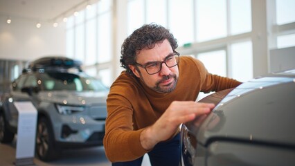 Camera view of attractive Caucasian man inspecting car carefully in auto salon. Customer looking...