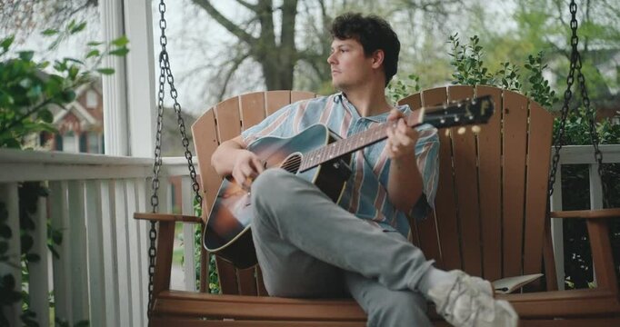 Young male musician sitting on a porch, playing guitar and writing a song
