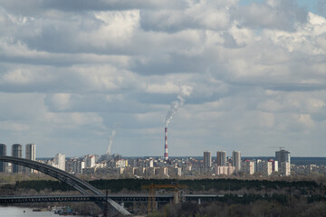 A sprawling city skyline features numerous high-rise buildings while smokestacks release smoke into the air. The scene captures the contrast of urban life against a cloudy backdrop