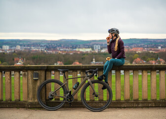 Blonde cyclist sits on stone fence, eating bretzel and gazing at panorama of city of Dresden in...