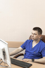 A male doctor in a blue suit sits at a computer and looks at the monitor