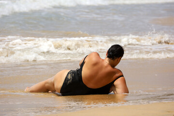 Woman in black swimsuit sunbathes lying on a sandy beach on sea waves background