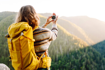 Rear view of young female traveler holding compass standing on cliff. Woman with hiking backpack...