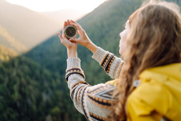 Rear view of young female traveler holding compass standing on cliff. Woman with hiking backpack...