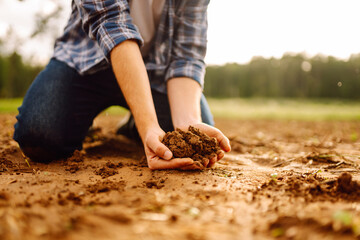 Close-up of the hands of a young agronomist in a checkered shirt holding fertile soil in an...