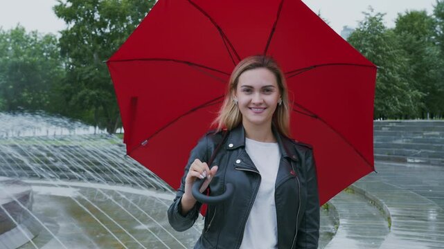 Blonde woman wearing leather jacket, standing near park fountain with red umbrella, radiating cheerful joy while smiling and looking at camera during rainy weather