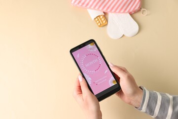 Menstruation. Woman holding smartphone with period calendar application on beige background, top...