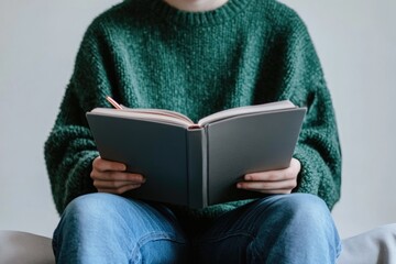 Person in green sweater sits reading a book and holding a pen.