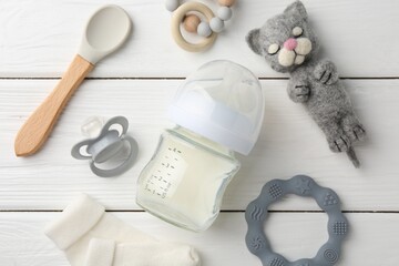 Feeding bottle with milk, needle felted cat, pacifier, spoon, baby socks and teethers on white wooden table, flat lay