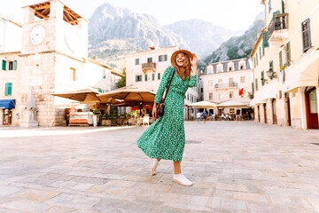 Naklejka premium A beautiful woman in a bright dress and hat walks through the old streets of Europe on a sunny day. A young tourist enjoys the sunrise outdoors. Tourism concept. Active lifestyle.
