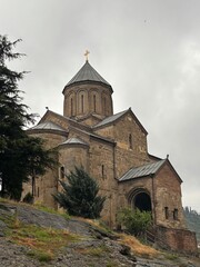 Fototapeta premium Ancient stone church on hill in Tbilisi surrounded by trees and cloudy sky