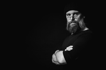 Fototapeta premium Black and white portrait of a stylish confident mature man in a black knitted hat with a beard and glasses. A keen look, an emotional gray beard and a black vest. A stylish restrained portrait on a da