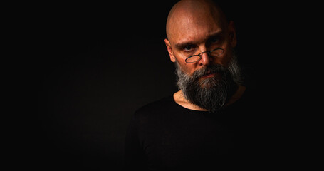 Bald bearded man with intense gaze and round glasses, half-lit in dramatic low key lighting. Mysterious look, wise expression, dark moody portrait