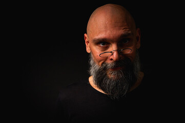 Bald bearded man with intense gaze and round glasses, half-lit in dramatic low key lighting. Mysterious look, wise expression, dark moody portrait