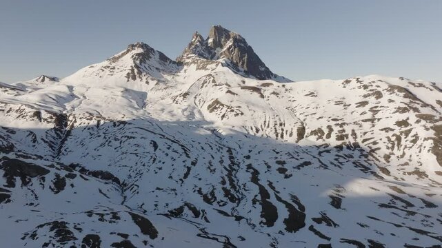 Aerial shot - Bright sunlight casting golden rays across snow blanketed slopes, revealing jagged Pyrenees peaks with iconic Midi d'Ossau standing against shared Franco Spanish mountain landscape