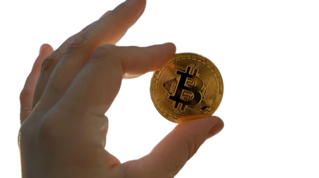 The image shows a close-up of a hand holding a physical representation of Bitcoin. The Bitcoin symbol "₿" in the center. You can place this background-removed PNG file wherever you like. - Powered by Adobe